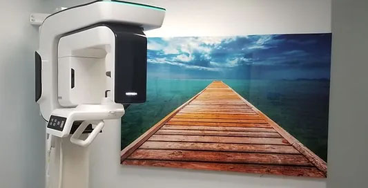 The image shows a modern dental or medical facility, featuring a large, high-resolution digital X-ray machine with a vibrant landscape photo in the background, and a sign with a wooden plank leading to a beach scene.