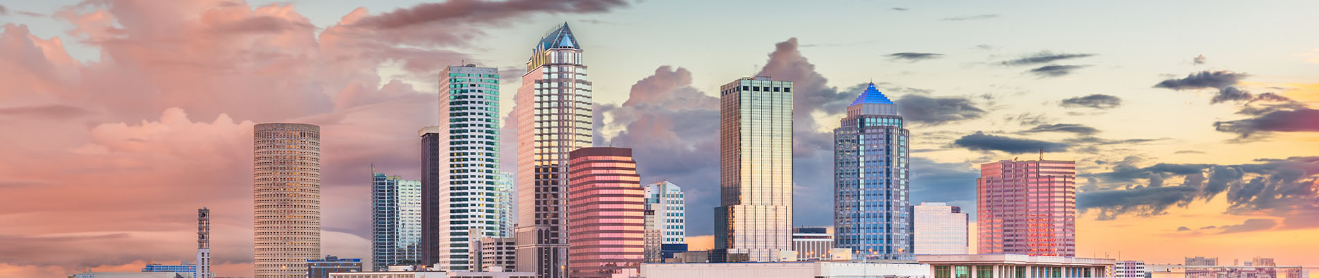 The image is a photograph of a city skyline at sunset, featuring a prominent downtown area with various buildings and a bridge silhouetted against the colorful sky.