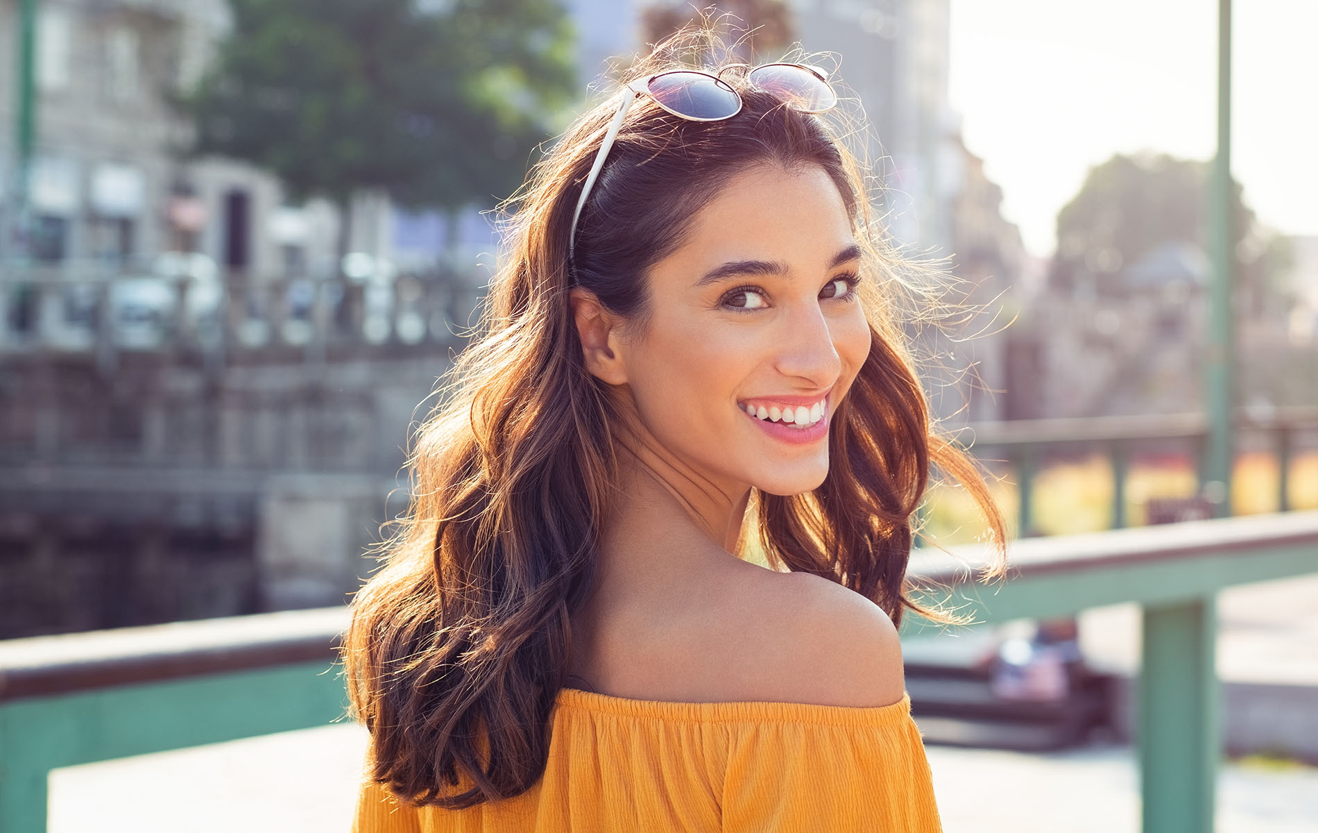 A young woman with a radiant smile, wearing a yellow off-the-shoulder top and sunglasses, stands against a backdrop of a cityscape.