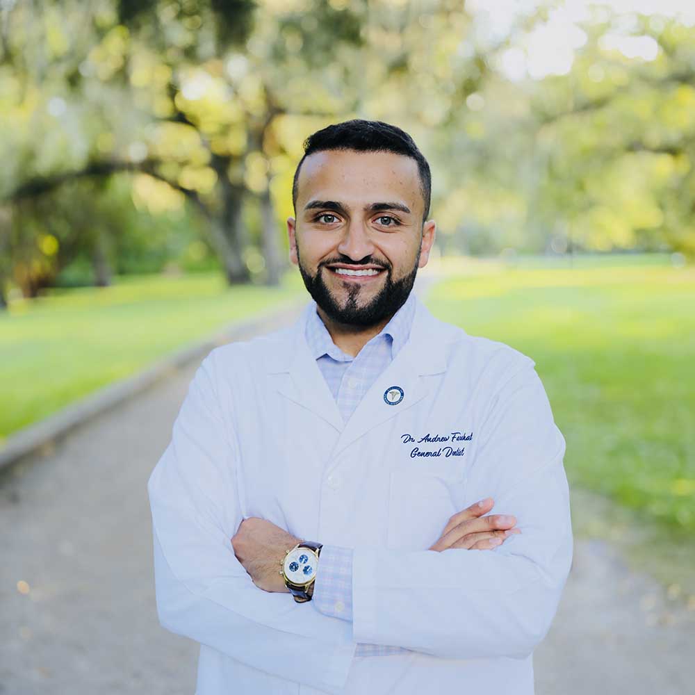 The image shows a man in a white lab coat, standing outdoors with a confident posture, smiling and posing for the camera.