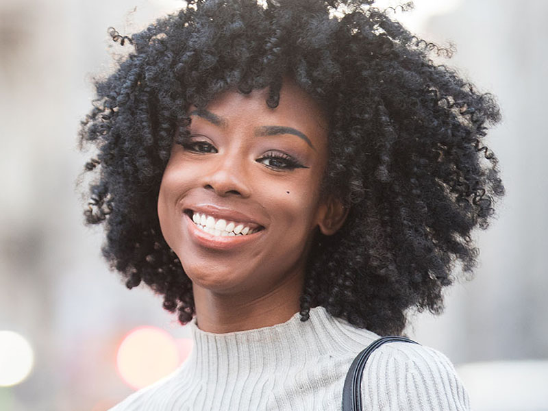 The image features a woman with curly hair, smiling and wearing makeup. She has a light complexion and is dressed in what appears to be casual attire. The background is blurred but suggests an urban setting with a building and possibly a street sign.